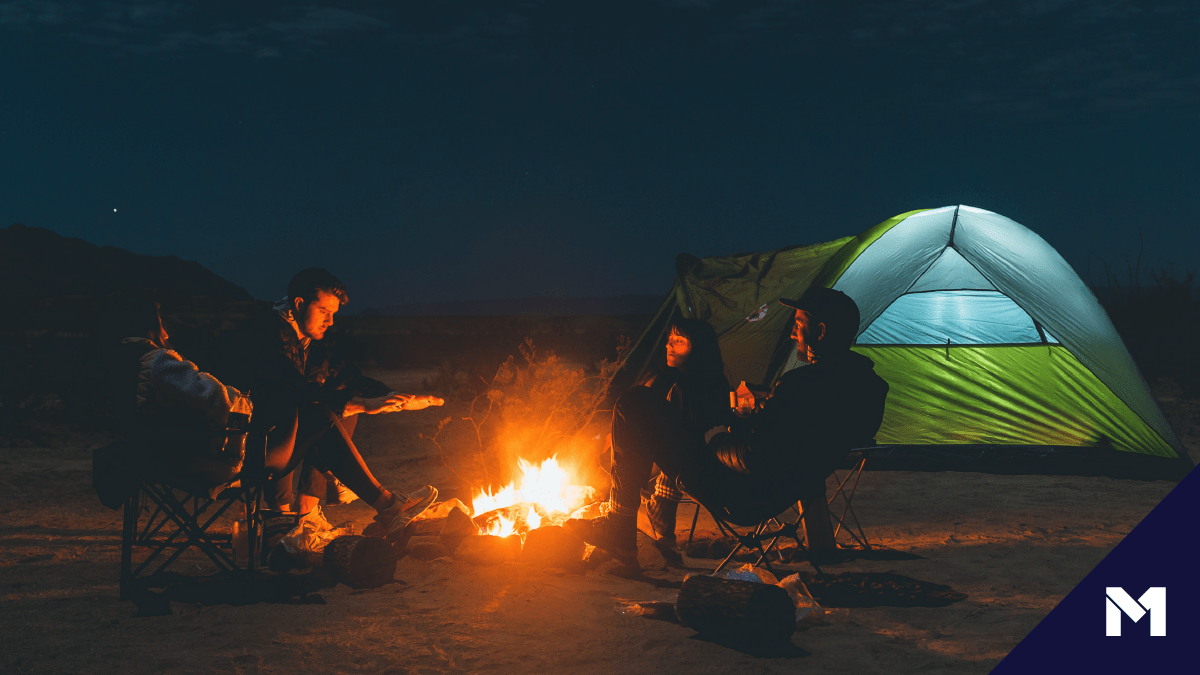 Image of three people around a campfire with a tent in the background and the M1 logo in the bottom right corner.