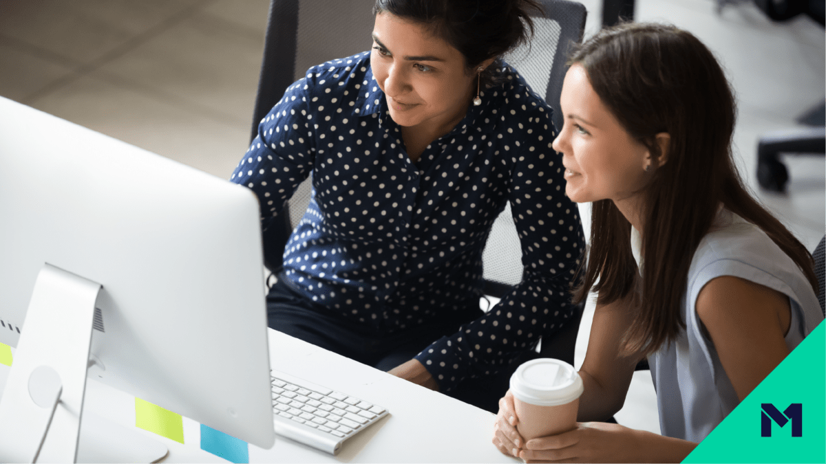 Two women seated in desk chairs smiling at a computer screen with the M1 logo in the bottom right corner