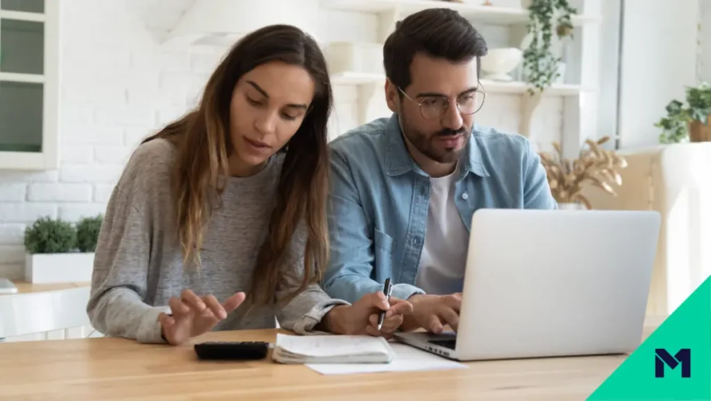 Couple sitting at table, with the woman performing calculations and the man researching on a laptop computer