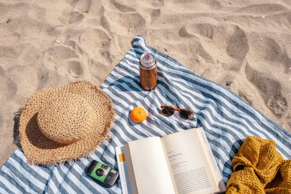 Beach towel with vacation essentials: a book, food, camera, straw hat, and drink